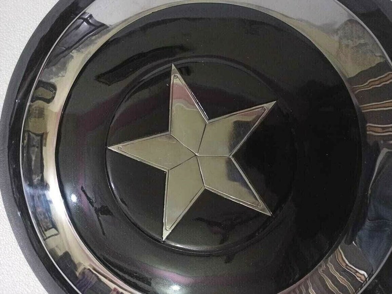 Puede incluir: Un escudo circular negro y plateado con una estrella de cinco puntas en el centro. La estrella es de plata y tiene una superficie reflectante.
