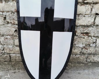 Puede incluir: Un escudo medieval con un diseño de cruz en blanco y negro. El escudo tiene un borde negro con remaches y una base puntiaguda. La cruz está centrada, con una franja vertical y una horizontal negras.
