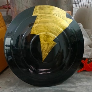 Op de afbeelding: Een rond zwart schild met een gouden bliksemontwerp. Het schild is gemaakt van hard materiaal en heeft een glanzende afwerking.
