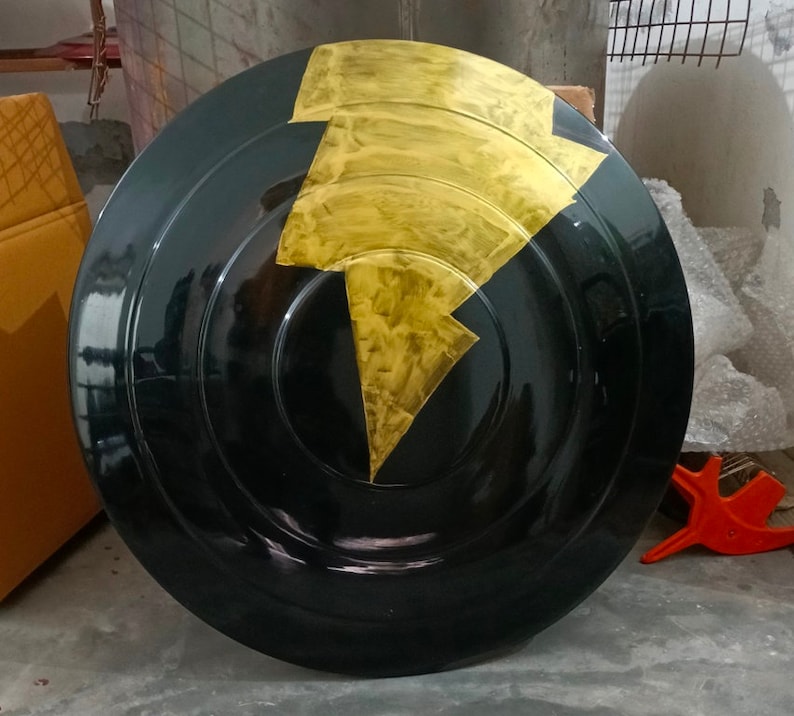 Op de afbeelding: Een rond zwart schild met een gouden bliksemontwerp. Het schild is gemaakt van hard materiaal en heeft een glanzende afwerking.