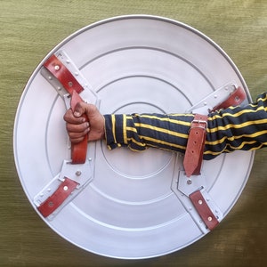 Op de afbeelding: Een rond, zilverkleurig schild met drie rode leren riemen. Het schild heeft een verhoogd midden en een cirkelvormig ontwerp. Een arm van een persoon houdt het schild vast.