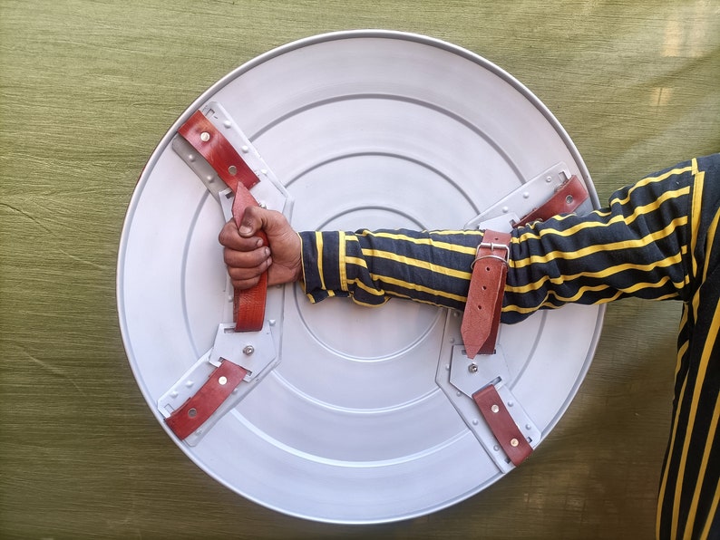 Op de afbeelding: Een rond, zilverkleurig schild met drie rode leren riemen. Het schild heeft een verhoogd midden en een cirkelvormig ontwerp. Een arm van een persoon houdt het schild vast.