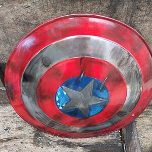 May include: A red, white and blue Captain America shield with a distressed, weathered look. The shield has a five-pointed star in the centre.