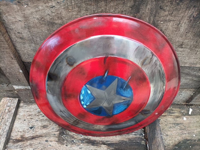 May include: A red, white and blue Captain America shield with a distressed, weathered look. The shield has a five-pointed star in the centre.