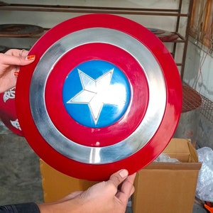 Op de afbeelding: Een rood, wit en blauw Captain America-schild met een zilveren metalen rand. Het schild wordt in de handen van een persoon gehouden.