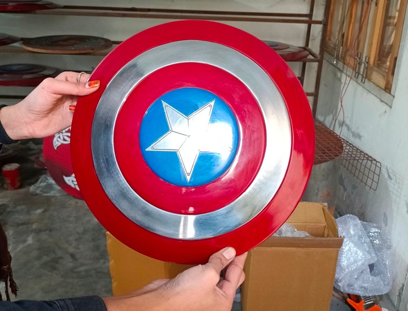Op de afbeelding: Een rood, wit en blauw Captain America-schild met een zilveren metalen rand. Het schild wordt in de handen van een persoon gehouden.
