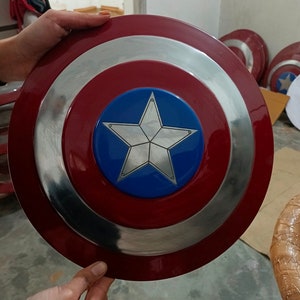 Op de afbeelding: Een rood, wit en blauw Captain America schild met een zilveren vijfpuntige ster in het midden. Het schild is gemaakt van metaal en heeft een glanzende afwerking.