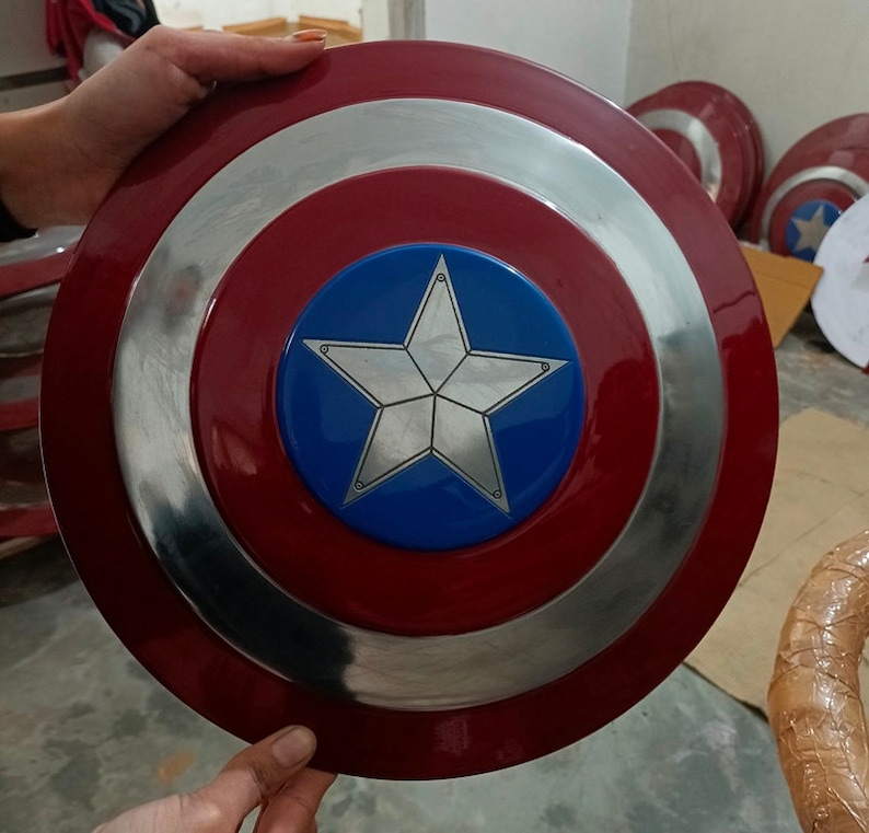 Op de afbeelding: Een rood, wit en blauw Captain America schild met een zilveren vijfpuntige ster in het midden. Het schild is gemaakt van metaal en heeft een glanzende afwerking.