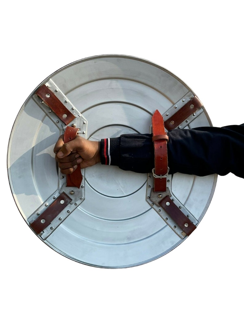 Op de afbeelding: Een rond zilveren schild met een rode leren riem en metalen klinknagels. Het schild heeft een verhoogd midden en wordt vastgehouden door een hand.