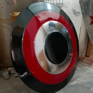 May include: A round shield with a black outer ring, a red middle ring, and a silver inner ring. The center is black. The shield is likely made of metal and has a glossy finish. It is a prop or decorative item.
