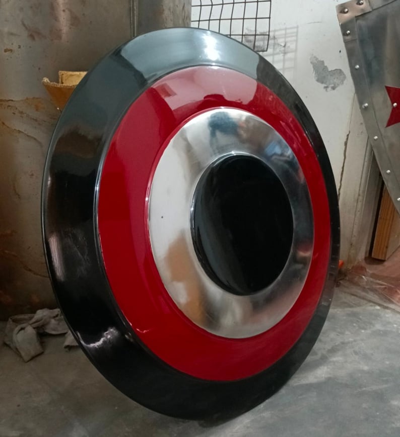 May include: A round shield with a black outer ring, a red middle ring, and a silver inner ring. The center is black. The shield is likely made of metal and has a glossy finish. It is a prop or decorative item.