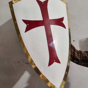 Puede incluir: Un escudo blanco con un diseño de cruz roja. El escudo tiene un borde de metal dorado y remaches. El escudo es un artículo histórico o decorativo.