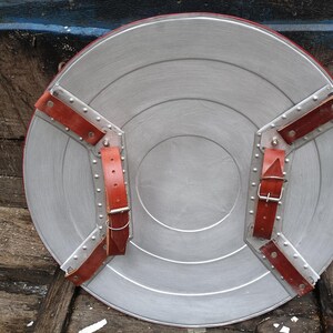 May include: A silver-colored, circular shield with a red border and brown leather straps. The shield has a series of concentric circles and is secured with riveted metal plates. The leather straps are buckled for secure handling.