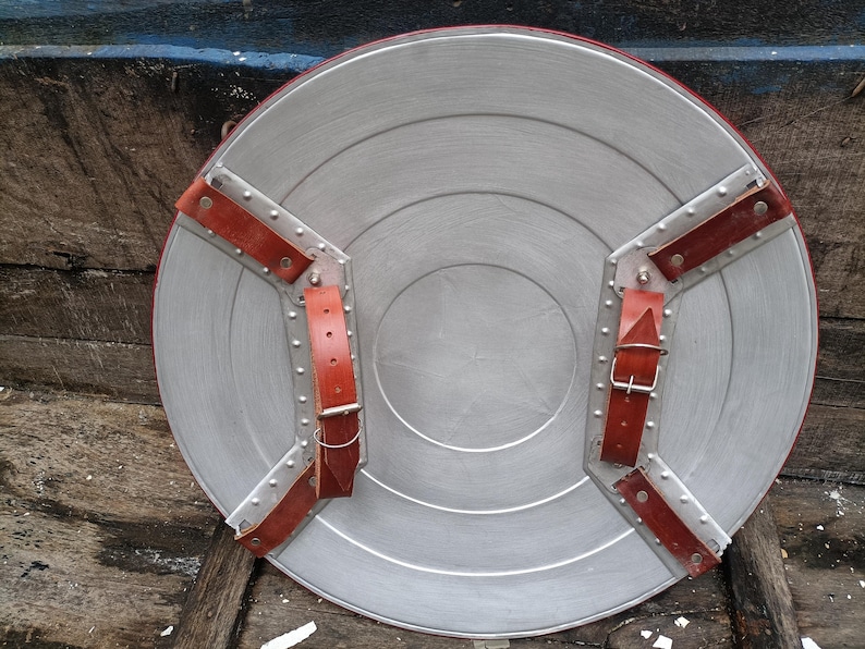 May include: A silver-colored, circular shield with a red border and brown leather straps. The shield has a series of concentric circles and is secured with riveted metal plates. The leather straps are buckled for secure handling.