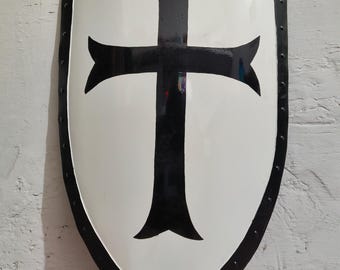Puede incluir: Un escudo blanco con un diseño de cruz negra. El escudo tiene un borde negro con remaches y una base puntiaguda. La cruz está centrada y tiene extremos ensanchados. El escudo es probablemente de metal.
