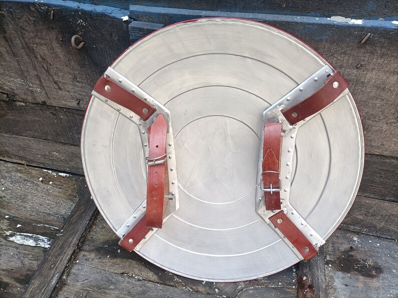 May include: A round, silver-coloured shield with two brown leather straps attached to the back. The straps have metal buckles and rivets.