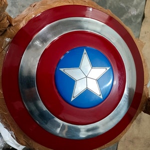 Op de afbeelding: Een rood, wit en blauw Captain America schild met een zilveren ster in het midden. Het schild is gemaakt van metaal en heeft een glanzende afwerking.