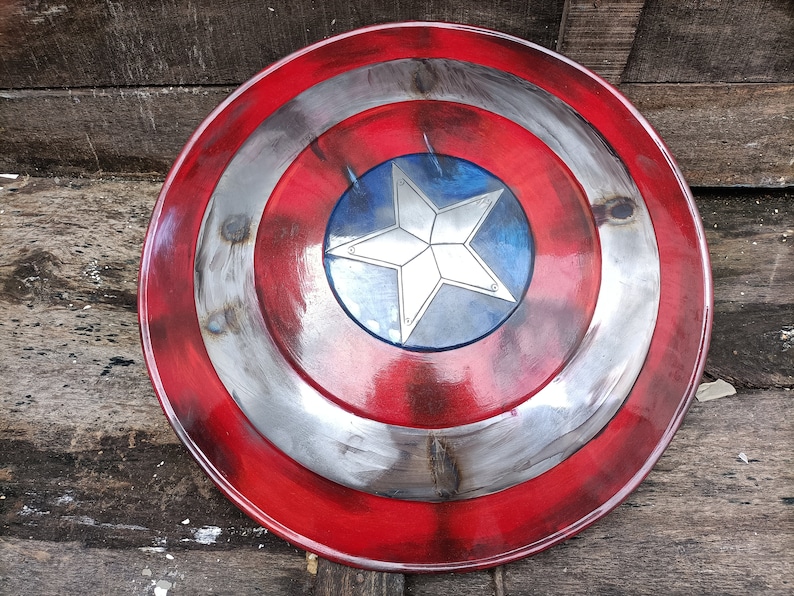 May include: A red, white and blue Captain America shield with a five-pointed star in the centre. The shield is made of metal and has a distressed look.
