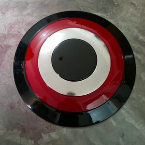May include: A round shield with a black outer ring, a red middle ring, a silver inner ring, and a black center. The shield has a glossy finish and is likely made of metal or a similar material.