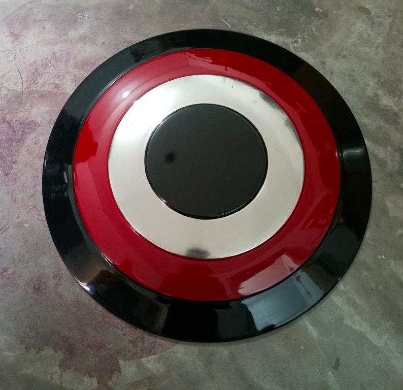 May include: A round shield with a black outer ring, a red middle ring, a silver inner ring, and a black center. The shield has a glossy finish and is likely made of metal or a similar material.