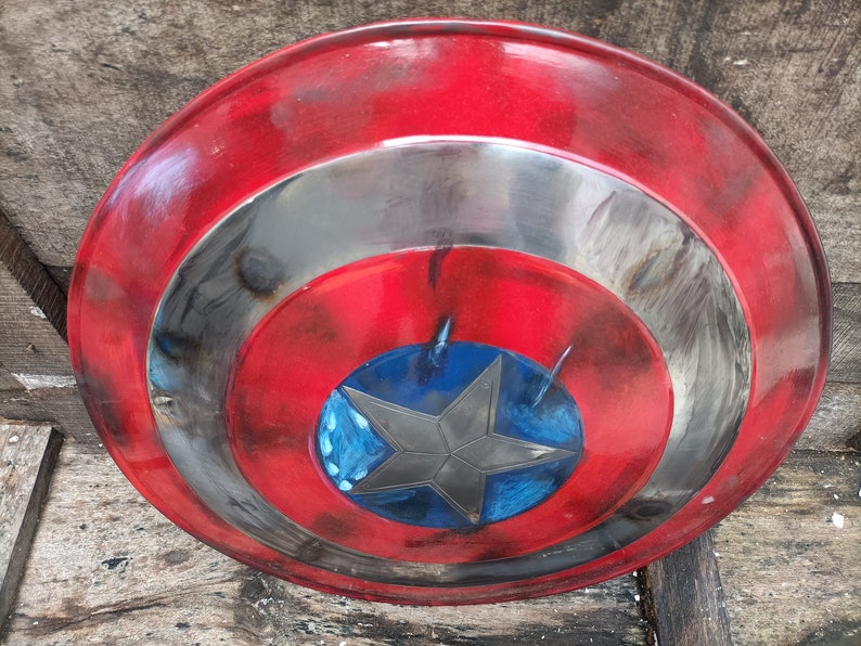 May include: A red, white and blue Captain America shield with a distressed paint finish. The shield has a five-pointed star in the centre.