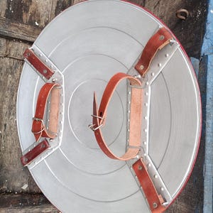 May include: A silver-colored, round shield with a red border and brown leather straps. The shield has a star-like design with rivets and is likely a costume accessory or collectible.