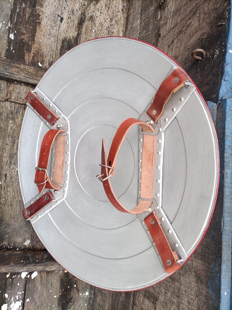 May include: A silver-colored, round shield with a red border and brown leather straps. The shield has a star-like design with rivets and is likely a costume accessory or collectible.