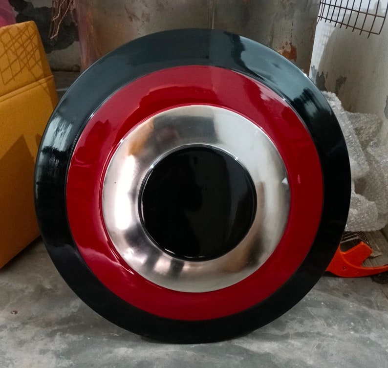 May include: A round shield with a black outer ring, a red middle ring, and a silver inner ring surrounding a black center. The shield has a glossy finish and appears to be made of metal.
