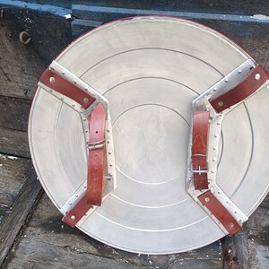 May include: A round, silver-coloured shield with two brown leather straps attached to the back. The straps have metal buckles and rivets.