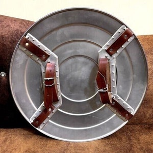Puede incluir: Un escudo redondo de color plateado con correas de cuero marr&oacute;n. El escudo tiene un centro elevado y un dise&ntilde;o circular alrededor del borde.