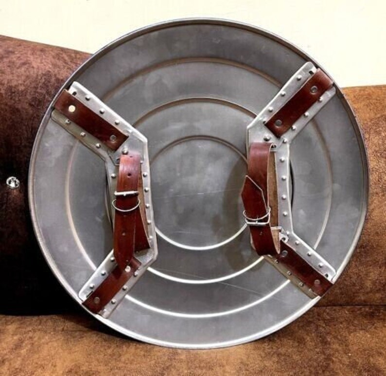 Puede incluir: Un escudo redondo de color plateado con correas de cuero marr&oacute;n. El escudo tiene un centro elevado y un dise&ntilde;o circular alrededor del borde.