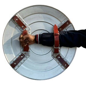 May include: A silver round shield with a red leather strap and metal rivets. The shield has a raised centre and is held by a hand.