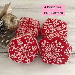 以下が含まれることがあります： 雪の結晶のデザインが施された、赤と白のビスコーヌのオーナメントが4つ。オーナメントは白い木製の表面に置かれています。画像の上部にはピンク色の文字で「4 Biscornu PDF Pattern」と書いてあります。