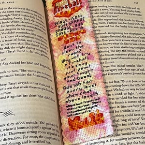 May include: A pink and orange bookmark with a cross-stitch design of a fireball spell. The bookmark includes text that reads "Fireball", "Range-150ft", "40ft Sphere", "Damage-B46", "Save-Dex 1/2", "Want to clean the room? Why not try a Fireball, it's easy, just point your finger and bam! Add 1d6 face every level when cast above level 3".