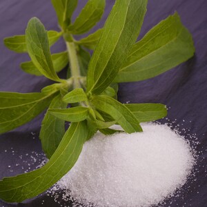 May include: A close-up shot of stevia leaves and a pile of white, granulated stevia sweetener. The green leaves are long and oval-shaped, with visible veins. The sweetener is a bright white color, contrasting with the dark background.