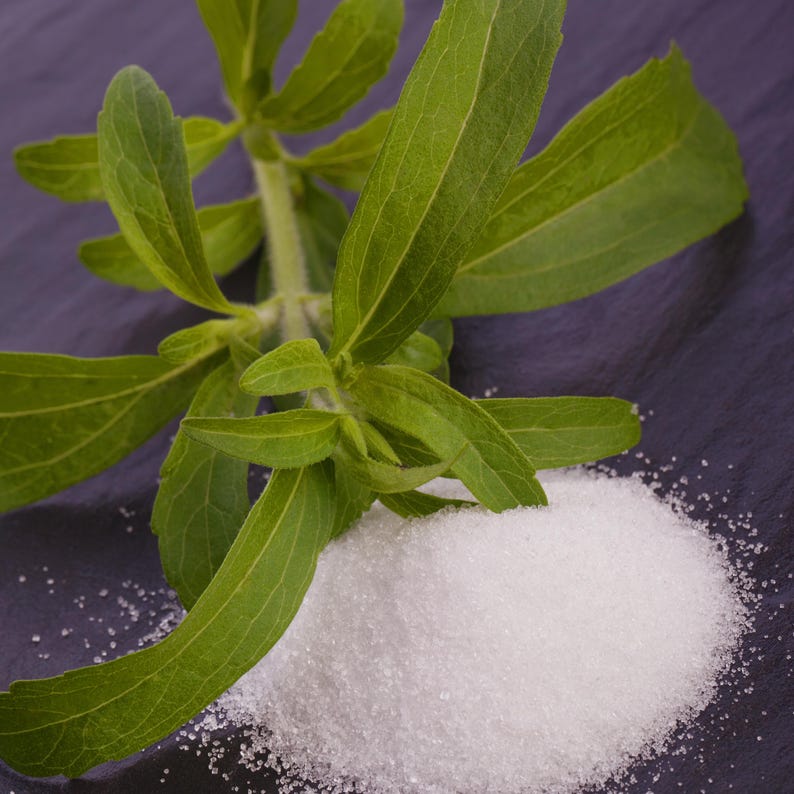 May include: A close-up shot of stevia leaves and a pile of white, granulated stevia sweetener. The green leaves are long and oval-shaped, with visible veins. The sweetener is a bright white color, contrasting with the dark background.