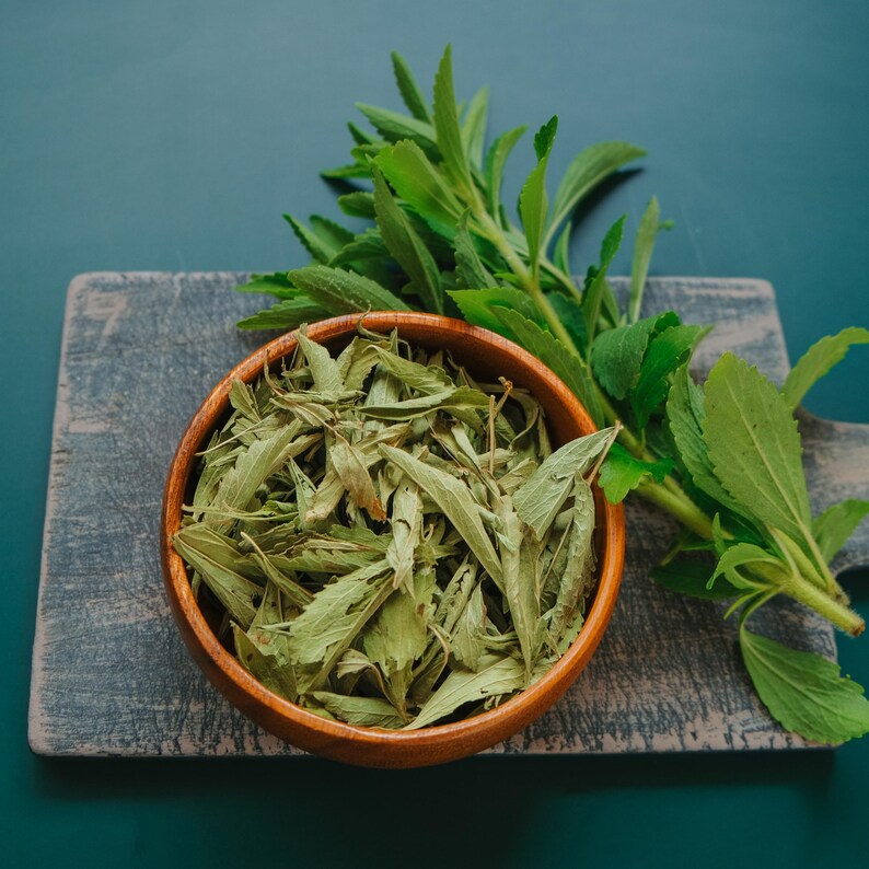 May include: A wooden bowl filled with dried green leaves, likely stevia, sits on a gray wooden cutting board. Fresh green stevia leaves are arranged to the right. The background is a dark teal color.