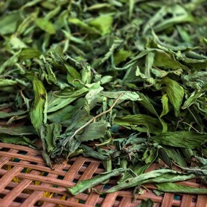 May include: Close-up of dried green stevia leaves, a natural sweetener. The leaves are a mix of light and dark green, with some showing signs of drying. The image is focused on the leaves, with a woven brown surface in the background.