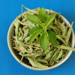 May include: A ceramic bowl filled with dried green stevia leaves and a fresh green stevia sprig. The bowl has a textured rim and sits on a bright blue surface. The image is a close-up shot.