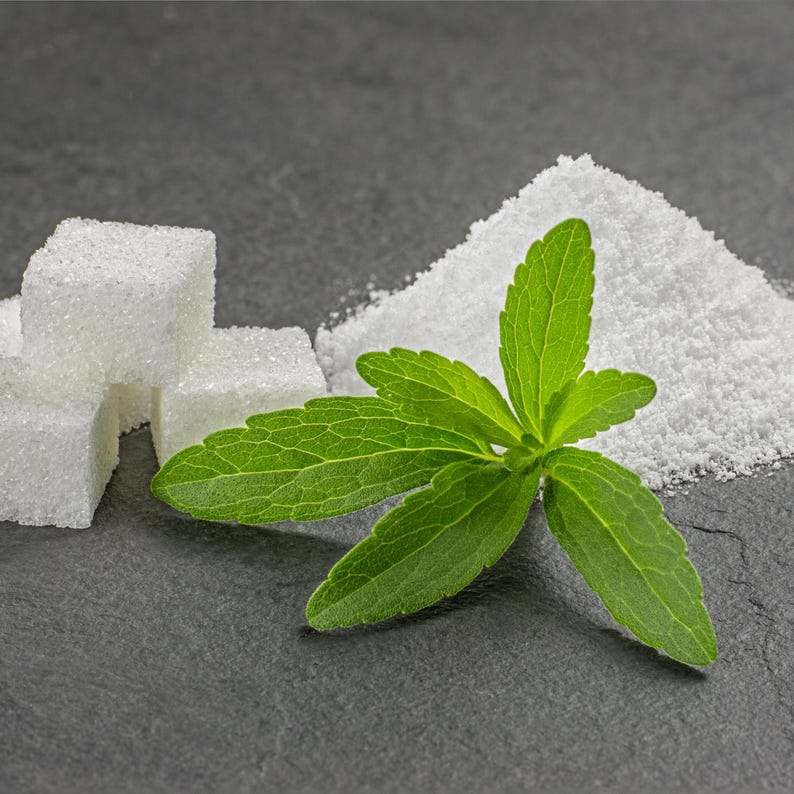 May include: A close-up shot features white sugar cubes, granulated sugar, and a sprig of green stevia leaves. The ingredients are arranged on a dark gray surface, highlighting the contrast between the white sugar and the green plant.