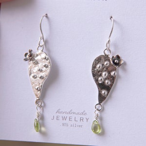 May include: A pair of handmade silver earrings. Each earring features a textured, leaf-shaped design with small, raised dots and a tiny flower at the top. A small, green peridot gemstone hangs from the bottom. The earrings are on a white card.