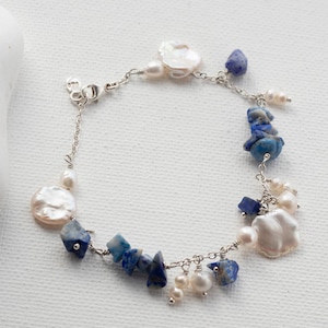 May include: A delicate bracelet featuring a silver chain adorned with blue lapis lazuli chips, white freshwater pearls, and irregular, flat, white pearl accents. The bracelet is displayed on a white surface.