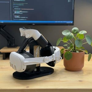 Könnte beinhalten: Ein weiß-schwarzes Virtual-Reality-Headset mit zwei passenden Controllern auf einem schwarzen Ständer mit gelbem Wabenmuster. Der Ständer verfügt über eine stabile ovale Basis und hält das Headset sowie die Controller in einer kompakten Anordnung.