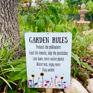 May include: A white metal sign with blue and white text that reads "GARDEN RULES" and lists rules for a garden, including protecting pollinators, feeding insects, skipping pesticides, using less lawn, planting more native plants, watering less, enjoying more, and staying awhile. The sign is surrounded by green plants and rocks.