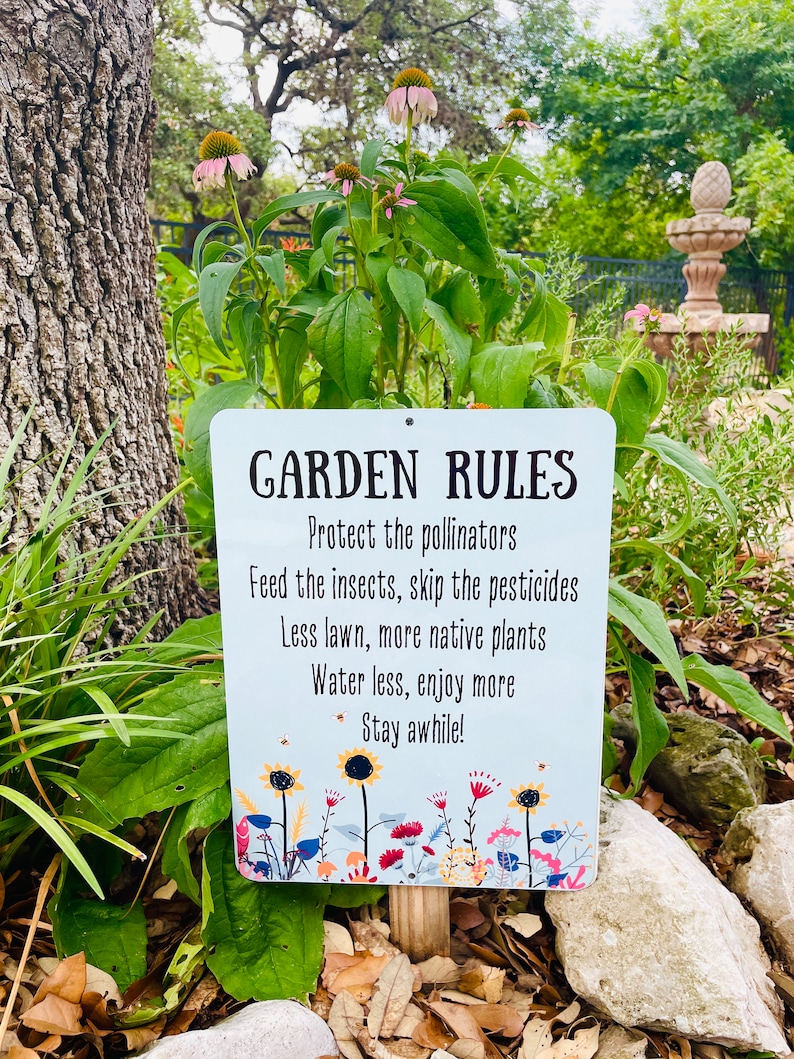 May include: A white metal sign with blue and white text that reads "GARDEN RULES" and lists rules for a garden, including protecting pollinators, feeding insects, skipping pesticides, using less lawn, planting more native plants, watering less, enjoying more, and staying awhile. The sign is surrounded by green plants and rocks.