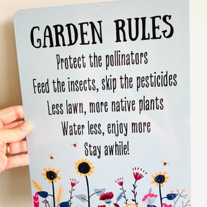 May include: A blue metal sign with white text that reads "GARDEN RULES" and a list of rules for gardening. The rules include protecting pollinators, feeding insects, skipping pesticides, using less lawn, planting more native plants, watering less, and enjoying more. The sign is decorated with a border of colorful flowers and bees.
