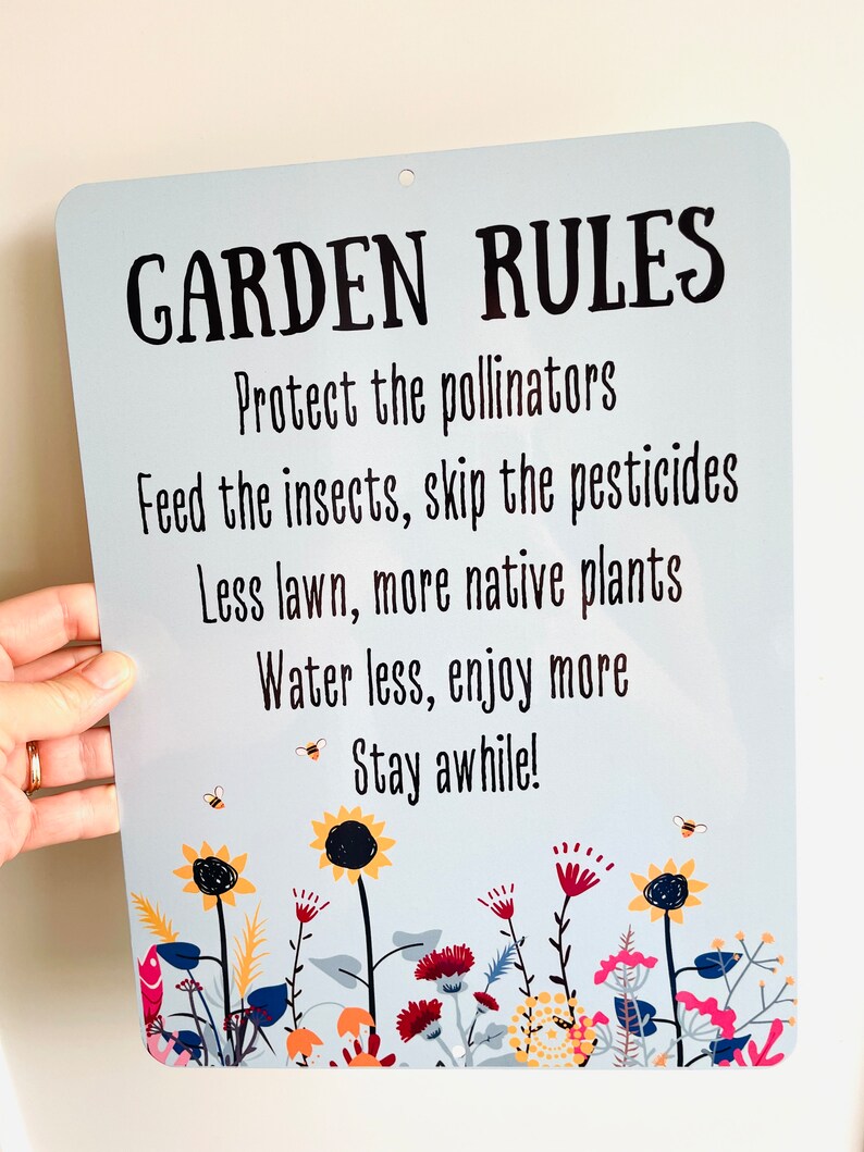 May include: A blue metal sign with white text that reads "GARDEN RULES" and a list of rules for gardening. The rules include protecting pollinators, feeding insects, skipping pesticides, using less lawn, planting more native plants, watering less, and enjoying more. The sign is decorated with a border of colorful flowers and bees.
