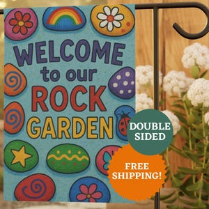 Puede incluir: Una bandera de jardín de doble cara con un fondo azul claro y diseños de rocas coloridas. La bandera dice "WELCOME to our ROCK GARDEN" en letras grandes. Las rocas decorativas incluyen un arcoíris, flores y una mariquita.