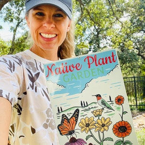 Może przedstawiać: Kobieta w szarej czapce z daszkiem z haftowanym napisem "Plant Natives" trzyma znak z napisem "Native Plant Garden". Na znaku widnieje koliber, motyl i kwiaty w otoczeniu ogrodu.