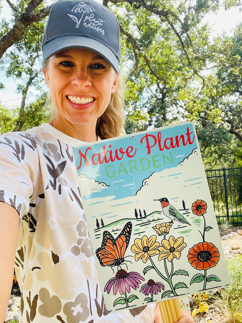 Może przedstawiać: Kobieta w szarej czapce z daszkiem z haftowanym napisem "Plant Natives" trzyma znak z napisem "Native Plant Garden". Na znaku widnieje koliber, motyl i kwiaty w otoczeniu ogrodu.